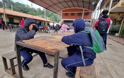 Estudiantes en uniforme azul sentados en bancos de madera bajo una estructura techada en lo que parece ser el patio de una escuela. En el fondo se observan edificios escolares con techos de tejas rojas y otros estudiantes. El ambiente sugiere un recreo o momento de descanso en una institución educativa.